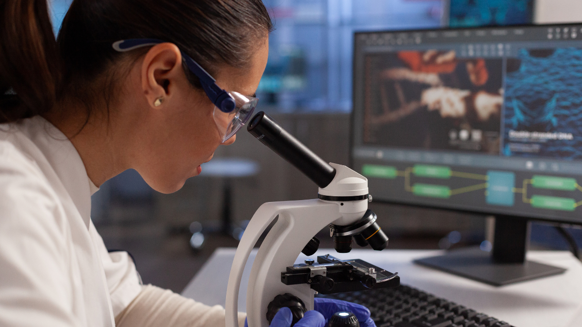 Pesquisadora em laboratório usando um microscópio e analisando uma amostra, com monitores ao fundo e interface de edição de imagem.