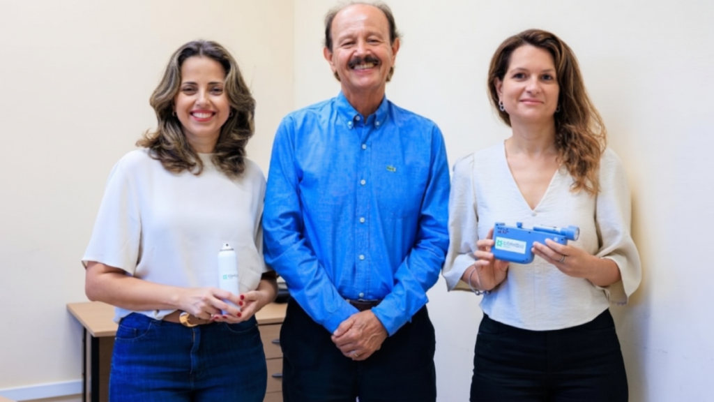 Três pesquisadores da Unicamp sorrindo em foto de equipe: uma mulher de blusa clara, um homem de camisa azul ao centro e outra mulher segurando um dispositivo azul, todos com postura profissional.