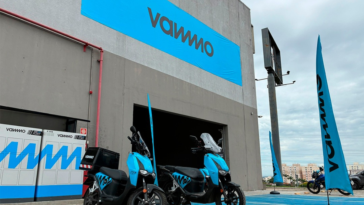 Fachada de empresa com letreiro VAMMO, área externa com motocicletas azul e estandartes verticais na entrada do showroom.