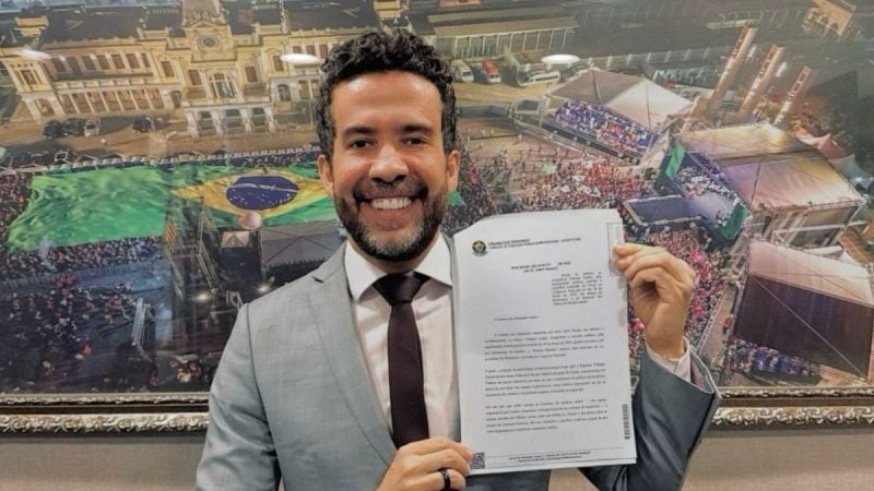 André Janones segura documento protocolado na Câmara dos Deputados, em frente a um quadro com imagem de manifestação.