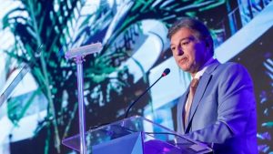 Claudio Lottenberg discursa em púlpito durante evento, usando terno azul e gravata, com painel digital ao fundo.