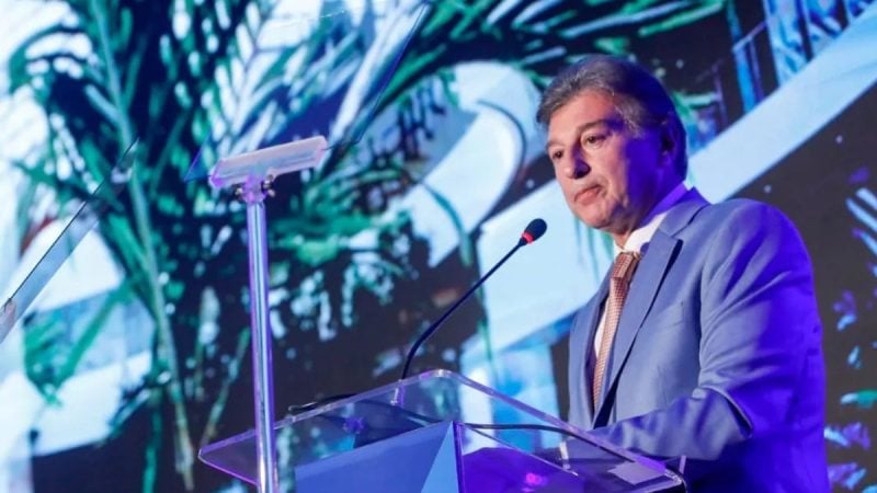 Claudio Lottenberg discursa em púlpito durante evento, usando terno azul e gravata, com painel digital ao fundo.
