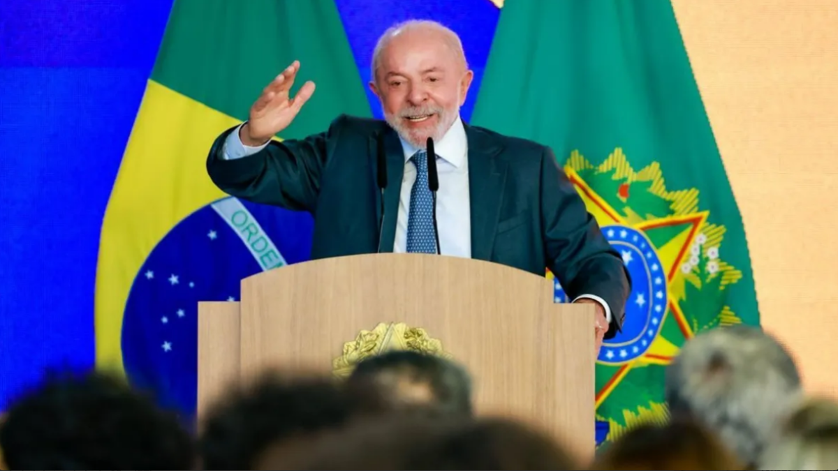 Imagem de Lula com terno escuro discursando em um púlpito, com bandeiras do Brasil ao fundo, em evento político.