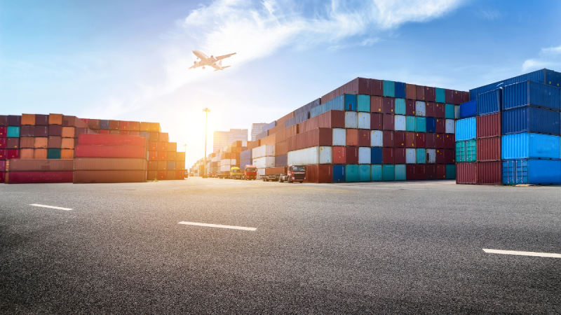 Pátio de contêineres de transporte com caminhões ao lado de pilhas coloridas, vista ampla de um porto e avião ao fundo no céu ensolarado