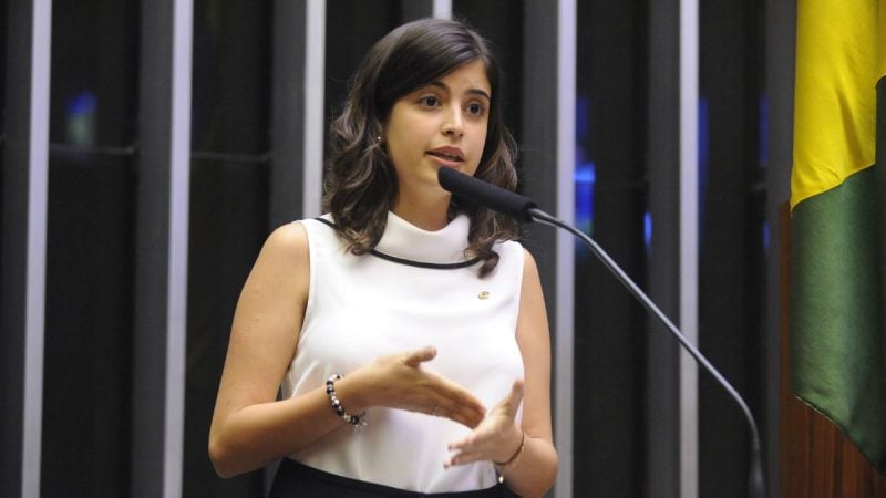 Deputada federal Tabata Amaral discursa ao microfone durante evento oficial, ao lado da bandeira do Brasil.