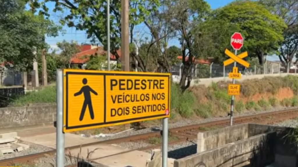 Placa de sinalização de pedestre na rodovia com a mensagem “PEDESTRE VEÍCULOS NOS DOIS SENTIDOS”, próximo a trilhos e vegetação ao fundo.