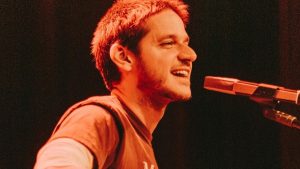 Cantor Rubel se apresenta ao vivo com violão e microfone durante show com iluminação avermelhada.