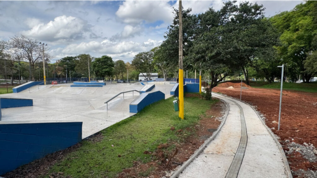 Parque infantil com pista de skate e área de playground, em Piracicaba com rampas, bancos e guarda-corpos azuis, caminho de concreto e área de terra preparada ao lado, sob céu nublado
