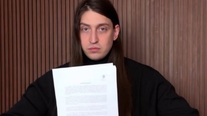 Felca segura um documento impresso enquanto posa sentado em uma poltrona, usando roupa preta.