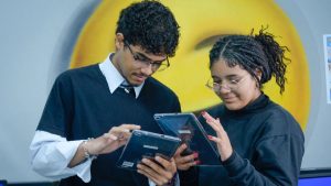 Dois jovens trabalhando juntos e consultando um tablet, com um homem apontando para uma tela e uma mulher concentrada, em ambiente de treinamento ou escritório.