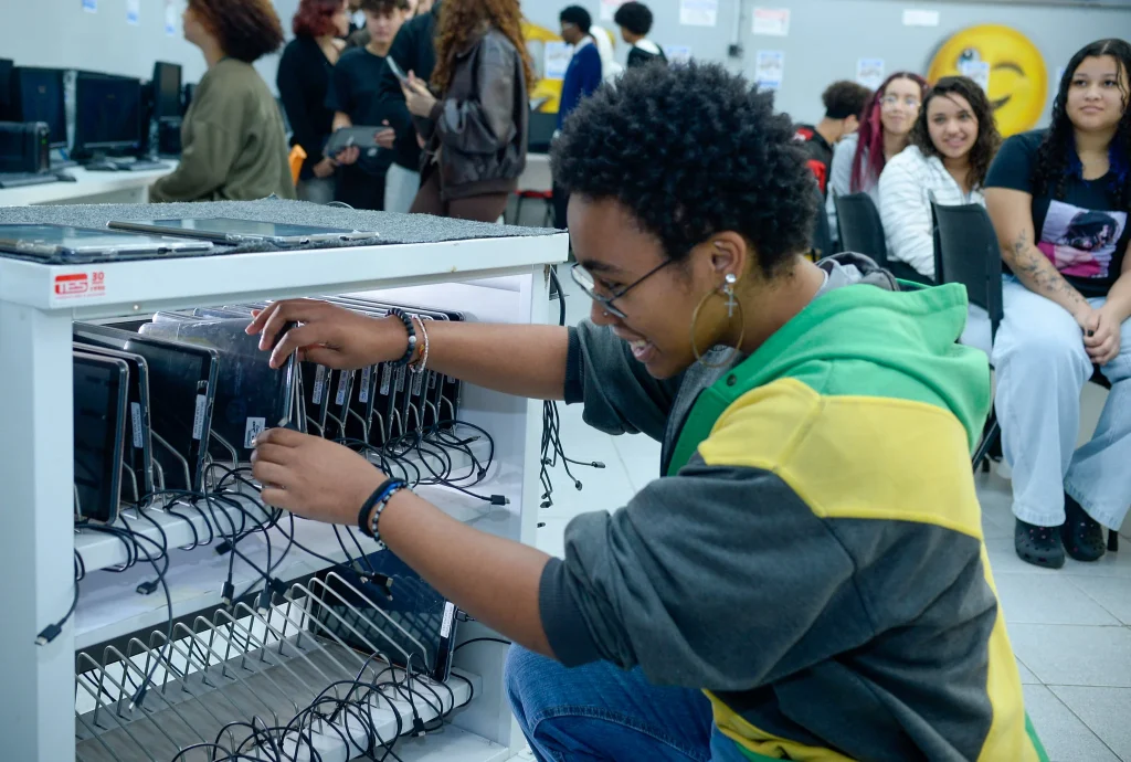 Aluna em escola estadual usando tablet em sala de informática, conectando e organizando os dispositivos enquanto outras pessoas observam ao fundo.