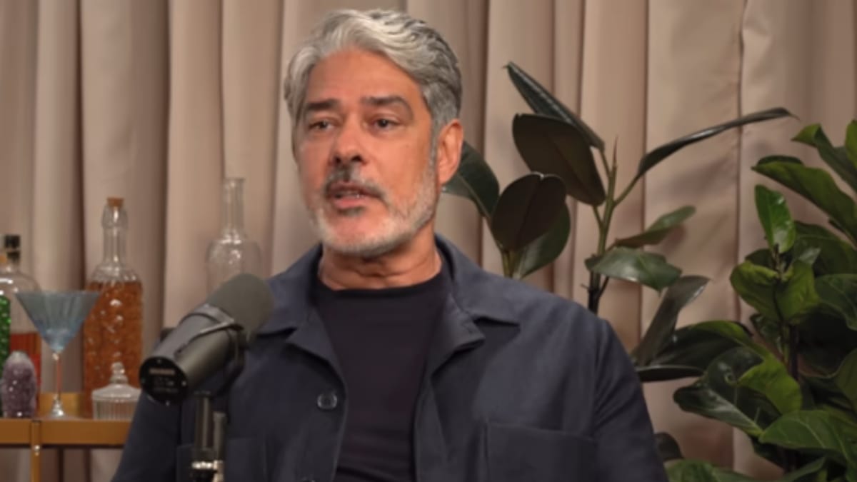 William Bonner em estúdio, falando ao lado de um microfone em ambiente interno com plantas ao fundo.