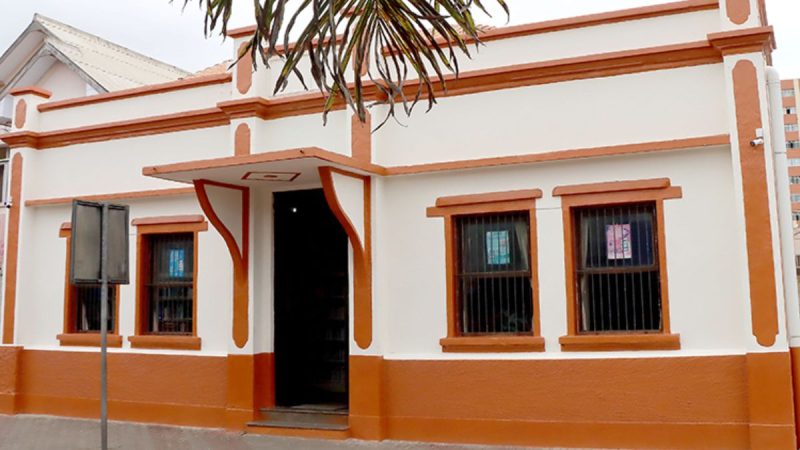 Fachada de casa térrea com paredes brancas e detalhes em laranja, janelas com grades e porta central com iluminação interna, vista externa do imóvel