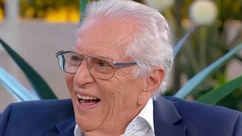 Carlos Alberto de Nóbrega sentado sorrindo. Apresentador usa óculos cinza, sorri e está com cabelos grisalhos