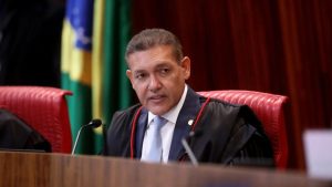 Imagem do presidente do TSE Kassio Nunes Marques sentado em uma cadeira presidencial de couro vermelho, em sessão solene, com microfone à frente e bandeiras ao fundo.