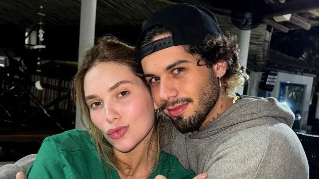 Casal abraçado em um ambiente interno, com uma mulher de cabelo claro e um homem de boné escuro, ambos sorrindo para a câmera.