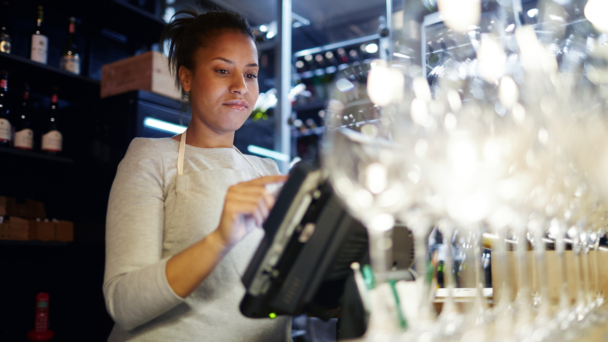Vendedora em uma adega bar iluminada, usando avental e atendendo clientes enquanto utiliza um tablet perto de prateleiras com garrafas de vinho. Foco no atendimento no ambiente de bebidas e iluminação noturna.