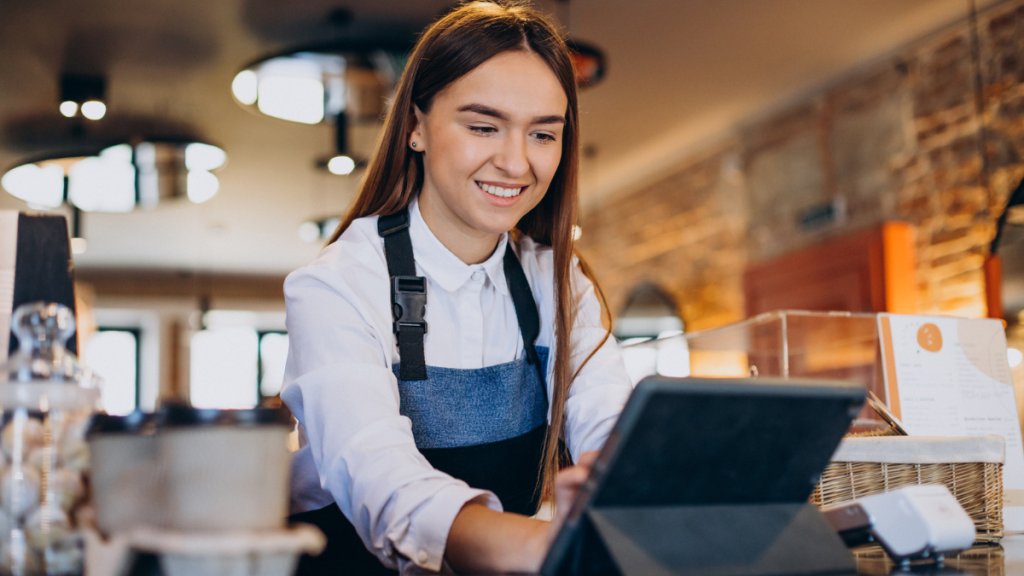 Mulher sorrindo como atendente em um café, usando avental azul e camisa branca, fazendo atendimento e olhando para um tablet em balcão com decoração de tijolos ao fundo.