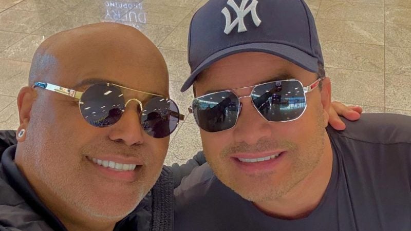 Rick e Renner sorriem para selfie em aeroporto, usando óculos escuros, enquanto posam juntos.