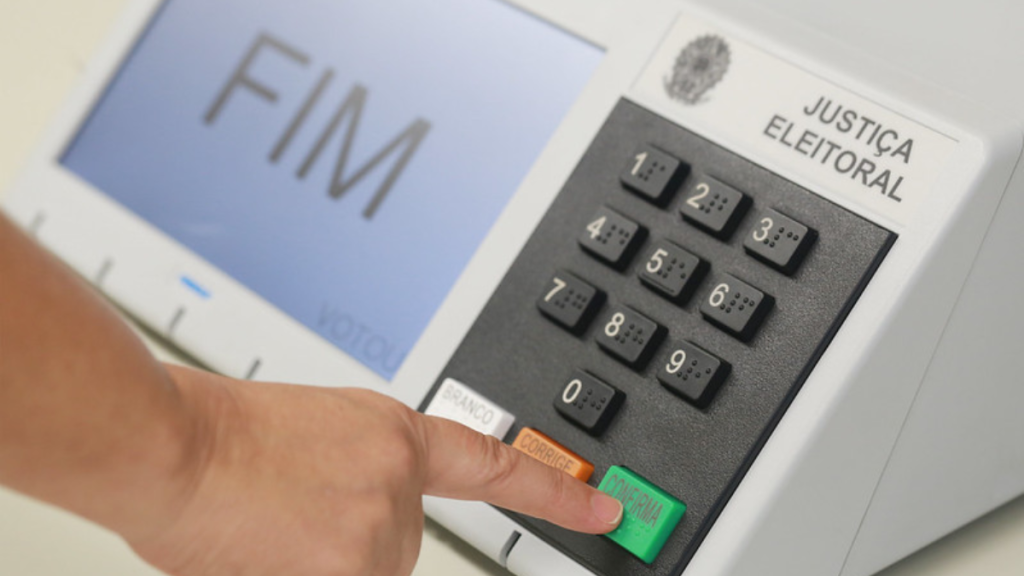 Detalhe da urna botão de seleção e dedos pressionando a opção “CORRIGE”, em um equipamento de caixa ou controle de justiça eleitoral.