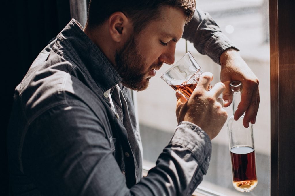 Homem demonstrando dependência de álcool, segurando uma taça com bebida alcoólica e fazendo movimentos de beber, enquanto observa a mesa ao lado em ambiente interno.