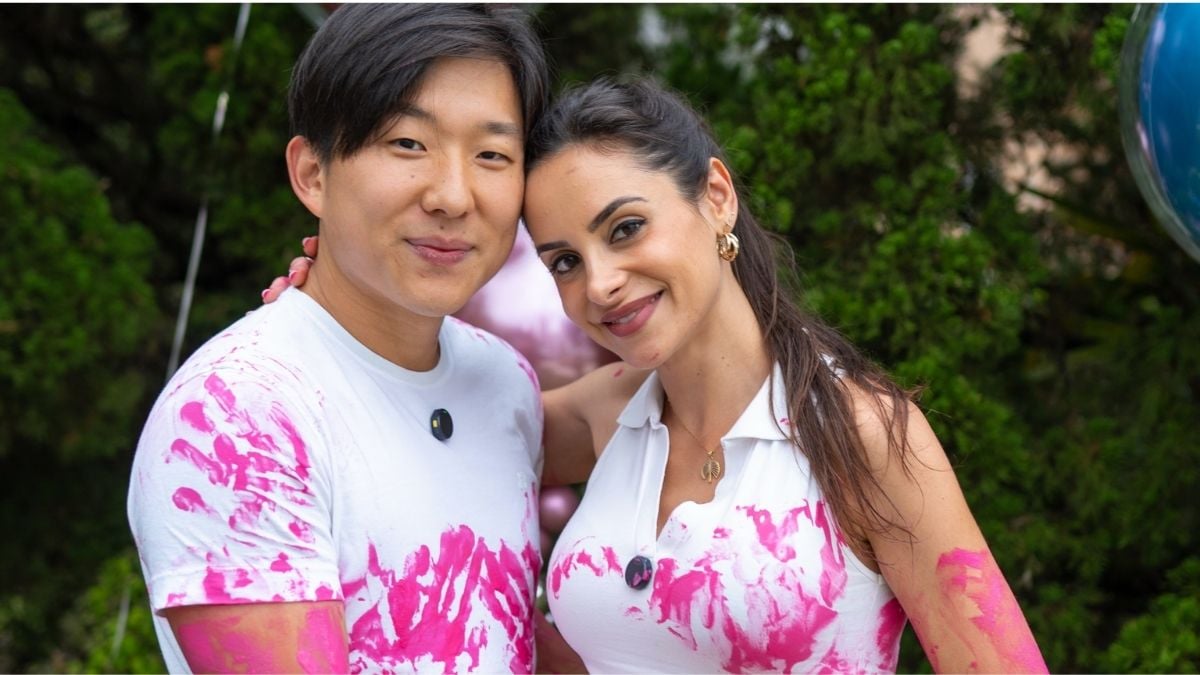 Pyong Lee e Natália Lee posam abraçados usando roupas brancas manchadas de tinta rosa em ambiente decorado com balões coloridos.