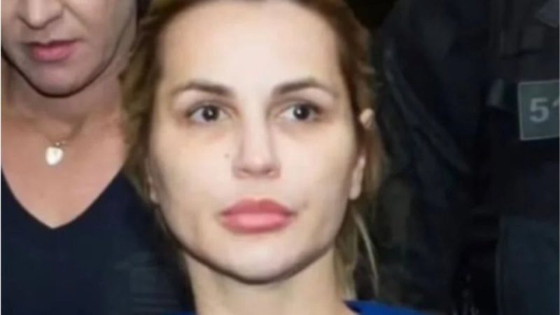 Primeiro plano da advogada e influenciadora Deolane Bezerra, de cabelo loiro e expressão séria, a ser escoltada por um agente da Polícia Civil.