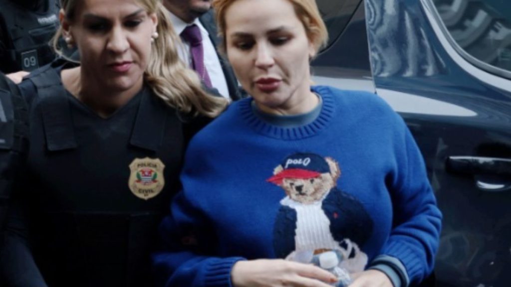 A advogada e influenciadora Deolane Bezerra vestindo um casaco azul com estampa de urso da Polo Ralph Lauren, escoltada por uma policial da Polícia Civil em frente a um veículo escuro durante operação policial.