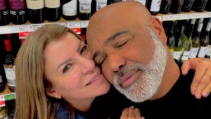 Geralda Helena e Rick Sollo em um momento de carinho, com rostos próximos e um beijo, em clima romântico em um ambiente com prateleiras ao fundo.