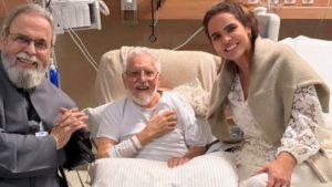 padre e Esposa e familiar visitam Carlos Alberto de Nobrega em leito hospitalar, com sorrisos e apoio durante o cuidado. Foto em ambiente de enfermaria.