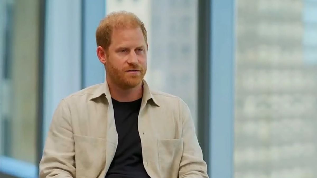 Príncipe Harry com barba curta usando camisa bege sobre camiseta preta, em ambiente interno com fundo desfocado, sentado e olhando para o lado durante entrevista ou conversa.