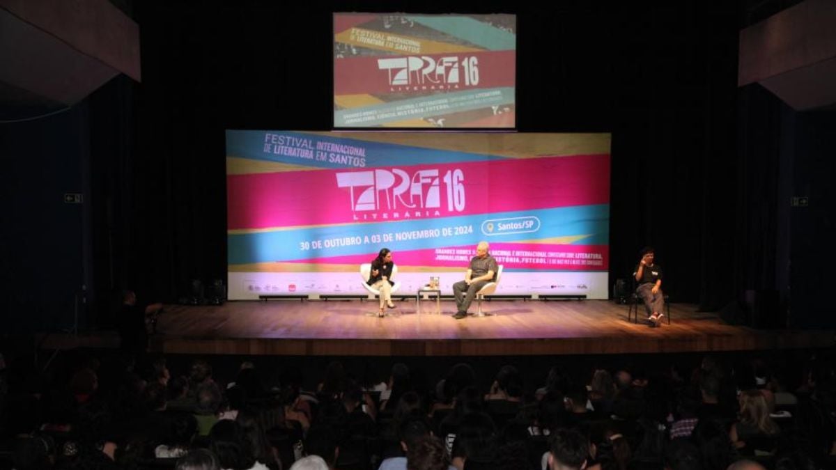 Palestra em palco durante o evento TRF 16, com dois palestrantes sentados em cadeiras e público assistindo em um auditório, telões exibindo programação da Tarrafa Literária em Santos/SP.