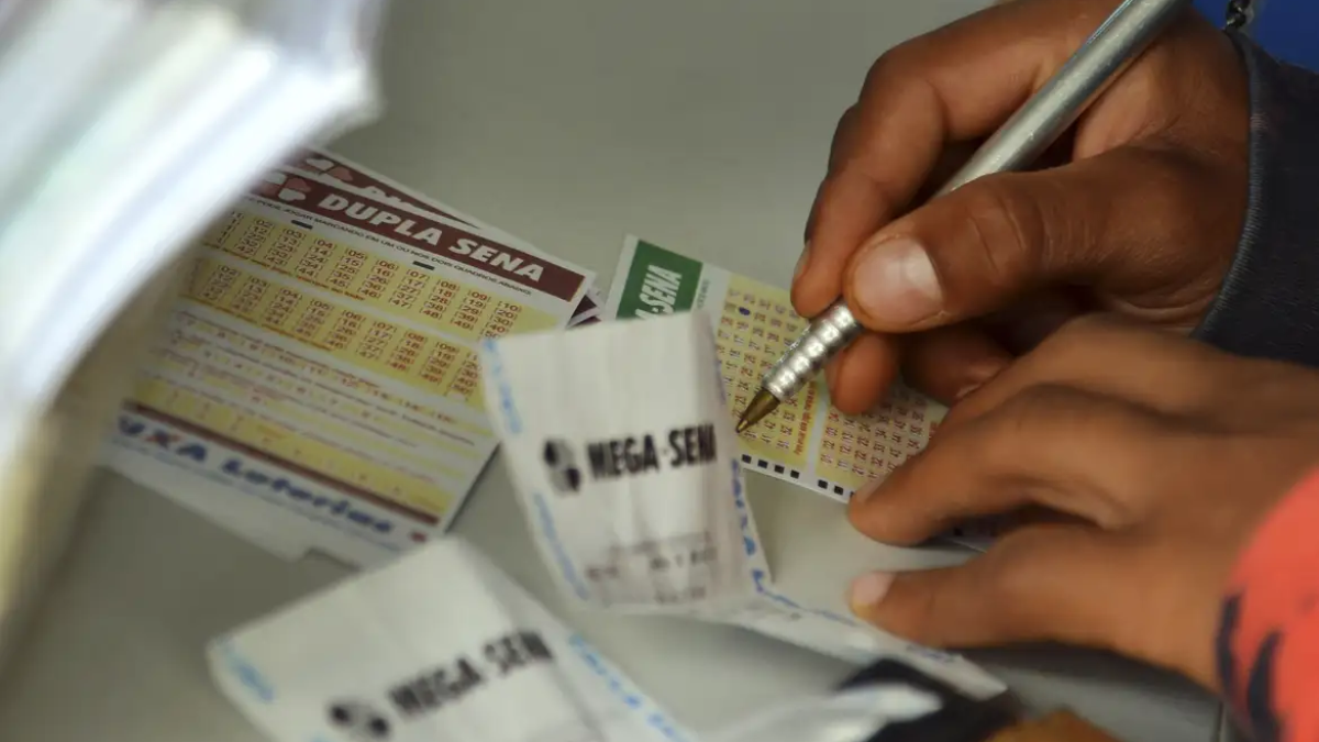 Pessoa preenchendo bilhetes de loteria com um caneta, enquanto vários jogos impressos estão sobre uma mesa; aparecem cartões com informações e colunas de aposta.
