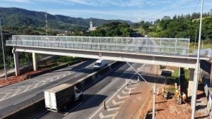 Passarela na Fernão Dias com estrutura metálica e proteção lateral sobre a rodovia, vista aérea com veículos ao fundo e equipes de obras em andamento.