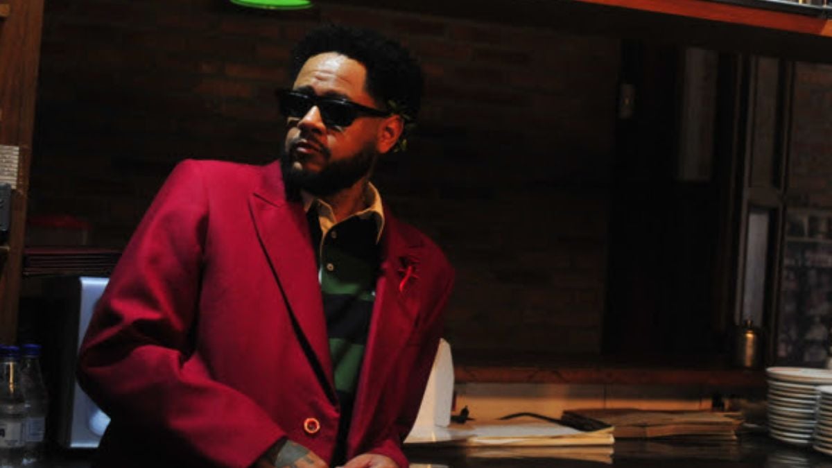 Emicida aparece em ambiente de estúdio, usando óculos escuros e blazer vermelho, com expressão confiante, diante de mesa com papéis e iluminação baixa