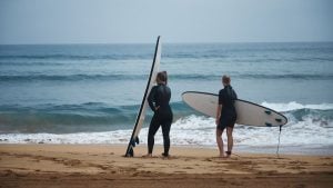 Dois surfistas em pé na areia de uma praia, segurando pranchas de surfe, com o mar ao fundo e ondas suaves se formando na costa.