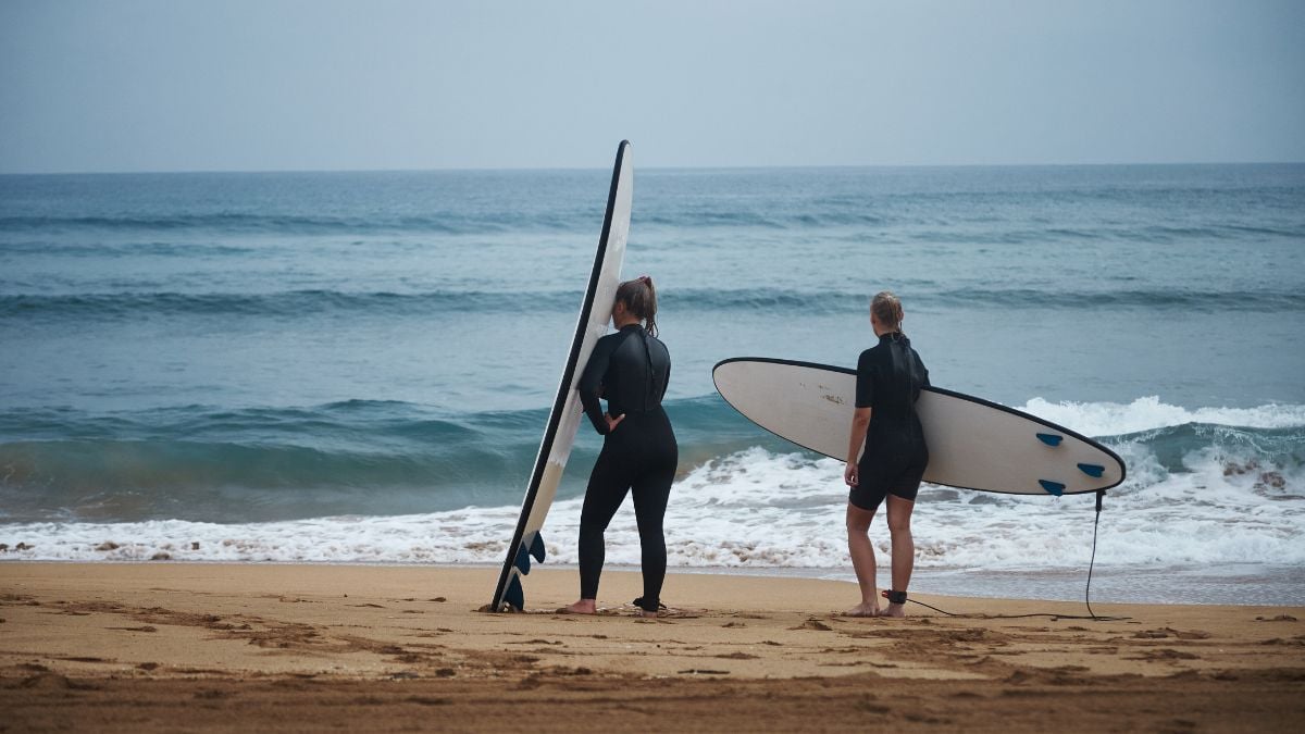 Dois surfistas em pé na areia de uma praia, segurando pranchas de surfe, com o mar ao fundo e ondas suaves se formando na costa.