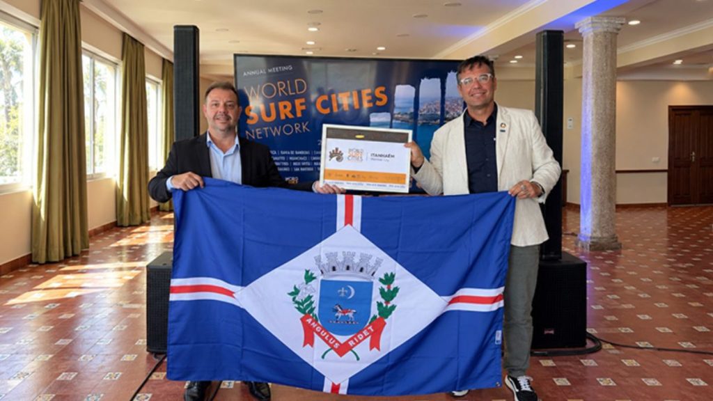 Dois homens em evento corporativo seguram um banner azul com o brasão do estado e a bandeira da região, em um salão com telão ao fundo que exibe o texto “World Surf Cities Network”.