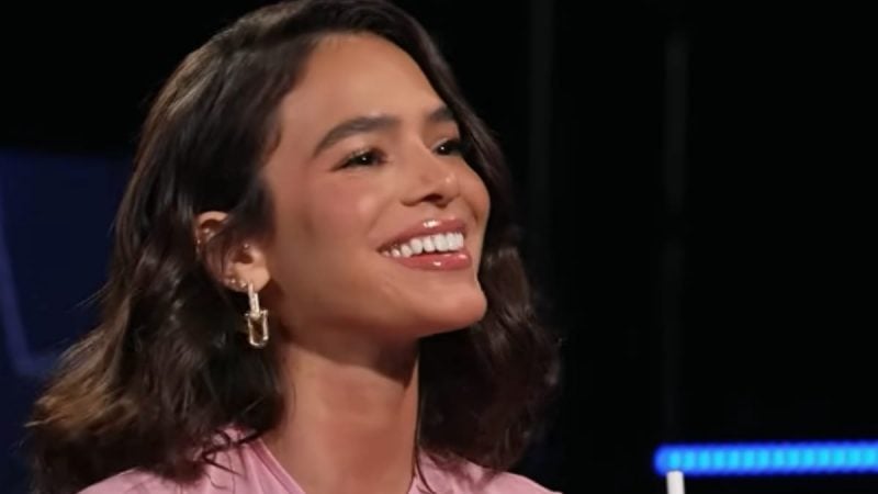 Bruna Marquezine em close, sorrindo no palco durante uma aparição em evento, com cabelo escuro ondulado e brincos pendentes, iluminação de ambiente.