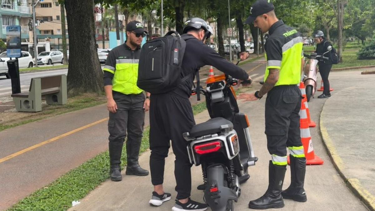 Equipe de técnicos uniformizados em via urbana, preparando e conferindo um robô para inspeção, com cones de sinalização e motocicletas ao fundo.