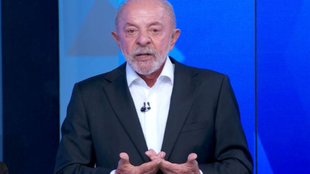 Luiz Inácio Lula da Silva fala durante o debate presidencial na televisão, vestindo terno preto e camisa branca, com as mãos erguidas em gesto explicativo contra um fundo azul de estúdio