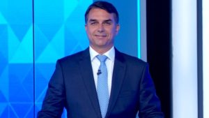 Flávio Bolsonaro sorri atrás de um púlpito em estúdio de televisão, vestindo terno azul-marinho, camisa branca e gravata azul-clara, com papéis de anotações apoiados à frente e painel azul ao fundo.