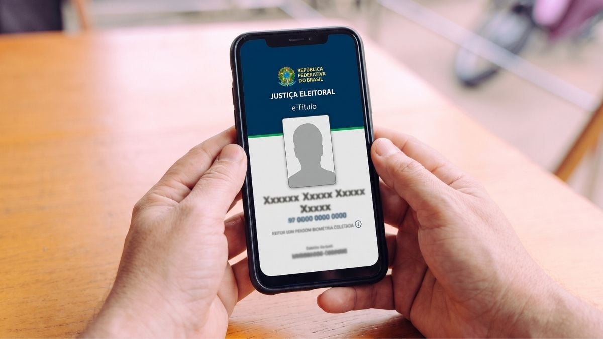 Mãos de um homem segurando um celular com o aplicativo do e-Título da Justiça Eleitoral aberto na tela sobre uma mesa de madeira.