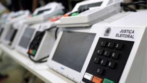 Fotografia em primeiro plano de várias urnas eletrónicas brancas da Justiça Eleitoral alinhadas sobre uma mesa, com o teclado numérico e o painel em destaque na primeira unidade e pessoas desfocadas ao fundo.