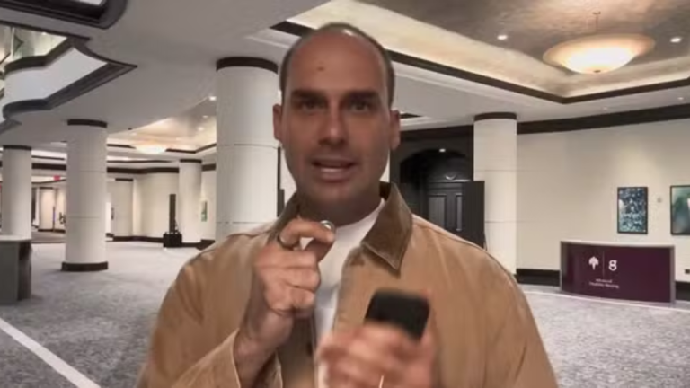 Eduardo Bolsonaro em um hall de hotel, falando com a câmera e apontando o dedo enquanto segura um celular na mão, em ambiente com colunas e iluminação no teto.
