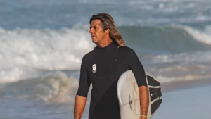 Dadá Figueiredo aparece carregando uma prancha, enquanto caminha pela praia.