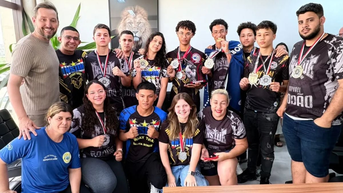 Equipe de atletas posa com várias medalhas e troféus durante premiação, em uma fotografia de grupo com pessoas de diferentes idades e uniformes esportivos