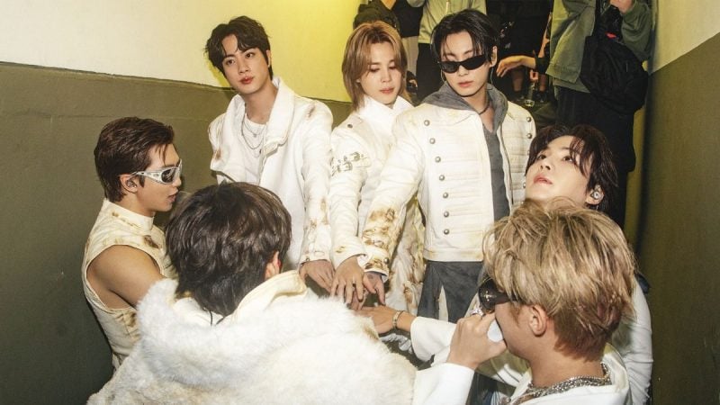 Foto do BTS com integrantes em roupas brancas, em um ambiente interno, posando juntos e encarando a câmera, em clima de bastidores