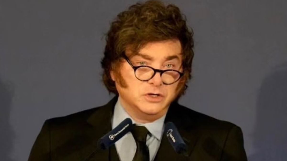 Javier Milei falando ao microfone, usando óculos e terno escuro com gravata, em um cenário de conferência ou entrevista.
