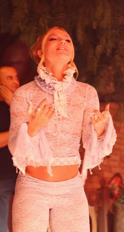 Virginia em foto artística usando roupa de renda branca com conjunto e detalhes delicados, em cena com iluminação quente e fundo desfocado.